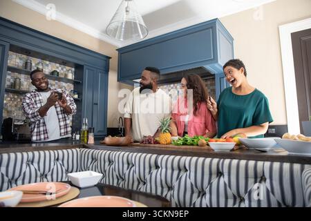 Diverse friends standing behind kitchen island, showing smartphone screen near pineapple and grapes Stock Photo