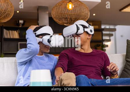 Diverse male friends wearing vr headsets, on sofa in living room with tumbler Stock Photo