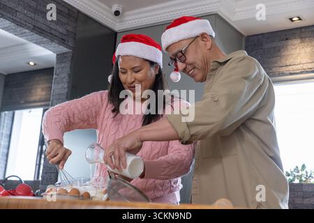 Diverse couple in santa hats whisking mixture while pouring milk into bowl at kitchen counter Stock Photo