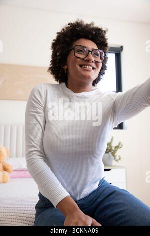 African American woman sitting on bed in bright bedroom wearing glasses and reaching for teddy bear Stock Photo