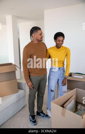 Diverse couple holding picture frame and bubble wrap standing among moving boxes in living room Stock Photo