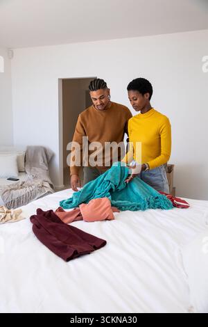 Couple folding teal blanket, burgundy sweater and peach shirt on white duvet in bedroom with sofa Stock Photo