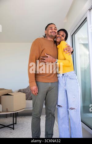 Diverse couple hugging and smiling in apartment with moving boxes side table sliding glass door rug Stock Photo