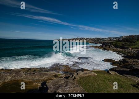 Experience the beauty of a serene coastal landscape featuring waves hitting rugged cliffs, under a vivid blue sky, with distant residential structures Stock Photo