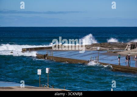 A seaside ocean pool with waves splashing on its perimeter, as individuals enjoy a relaxing walk near the water amidst scenic coastal views Stock Photo