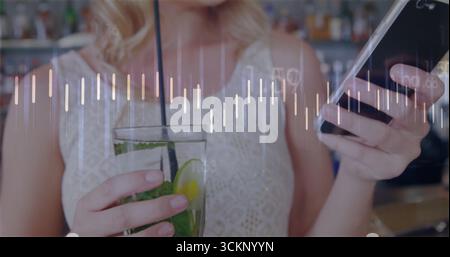 Woman wearing lace top holding tall cocktail and scrolling phone at bar counter, liquor bottles Stock Photo
