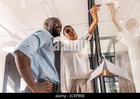 Diverse coworkers writing diagrams on glass board with printed paper, colored markers in office Stock Photo