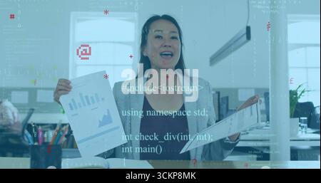 Presenting Korean woman wearing grey blazer displaying charts at office desk, with monitors Stock Photo