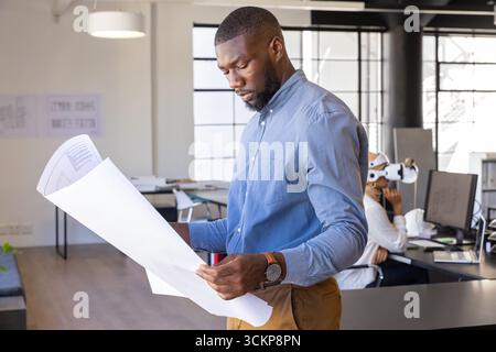 Diverse coworkers holding blueprints, using vr headset in modern open-plan office workspace Stock Photo
