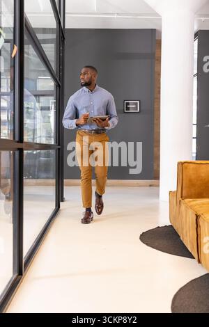 African american man walking in office corridor in business casual using stylus on digital tablet Stock Photo