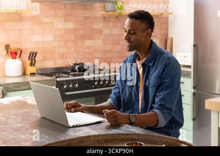Mid adult african american man working on silver laptop at kitchen island next to stove, copy space Stock Photo
