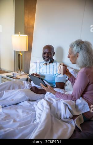 Diverse senior couple sitting up in bed holding mug and using tablet by nightstand with lamp Stock Photo