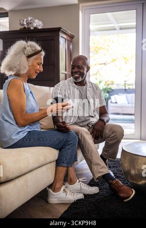 Diverse senior couple using digital blood pressure cuff on sofa in living room near sliding door Stock Photo