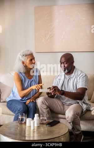 Diverse senior couple using weekly pill organizer on sofa at home with pill bottles and smartphone Stock Photo