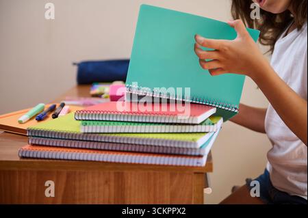 A student reviews several colorful spiral notebooks while sitting at a desk. Various writing instruments and materials are neatly arranged on the desk Stock Photo