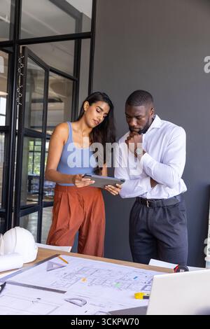 Diverse coworkers reviewing tablet with blueprints, ruler, helmet in modern office workspace Stock Photo