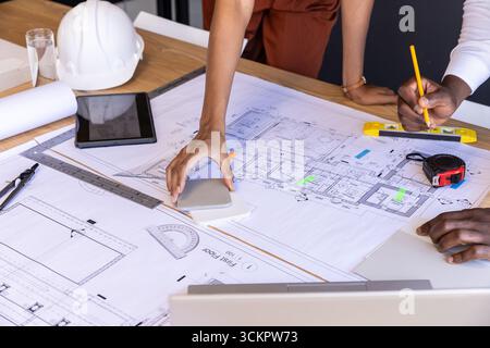 Diverse colleagues wearing rust jumpsuit marking blueprint on table in conference room Stock Photo