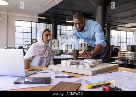Diverse coworkers examining architectural scale model at modern office with blueprints, laptop Stock Photo