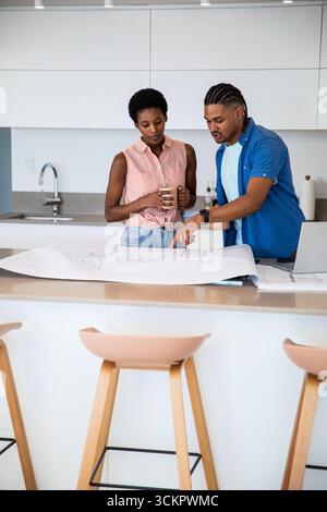 Diverse couple standing at kitchen island pointing at blueprint while holding ceramic mug Stock Photo