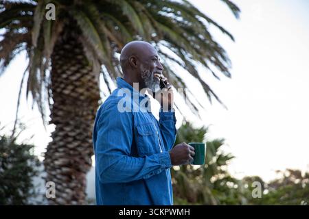Senior african american man standing in garden by palm fronds, calling on smartphone, green mug Stock Photo