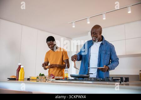 African american father and son cooking on kitchen island with spatula, stainless steel frying pan Stock Photo