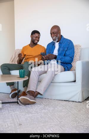 African american father and son sharing smartphone, sitting on sofa at home with green mug Stock Photo