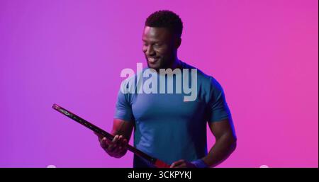 Smiling man wearing blue tee and wristband inspecting black racket under magenta lights in studio Stock Photo