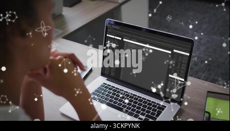 Reviewing mid adult woman examining terminal windows on laptop at workspace, with network overlays Stock Photo