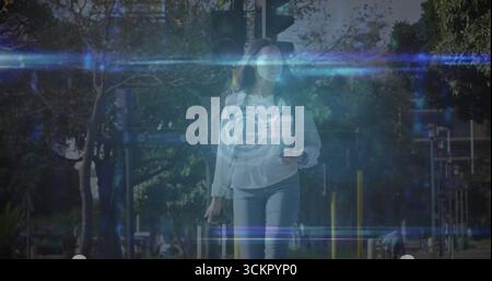 Walking Hispanic woman pulling wheeled suitcase, checking smartphone on sidewalk, traffic light Stock Photo