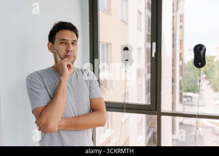 Thoughtful homeowner male contemplates looking at camera which robotic window cleaner to use, comparing white model with black one Stock Photo