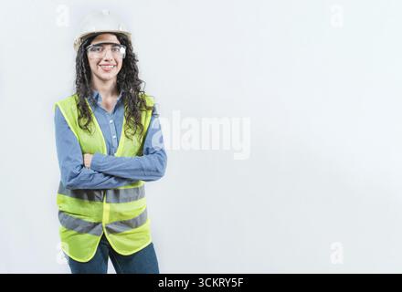 Portrait of smiling female engineer with arms crossed isolated. Young female engineer with helmet and vest looking at camera isolated Stock Photo
