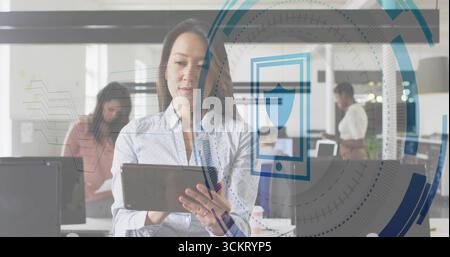 Interacting mid-adult woman holding tablet in open-plan office, with colleagues reviewing documents Stock Photo