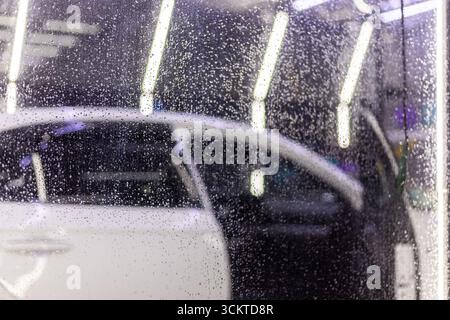 A white car is being washed in an indoor car wash detailing center behind wet plastic curtain. Close view with selective focus Stock Photo