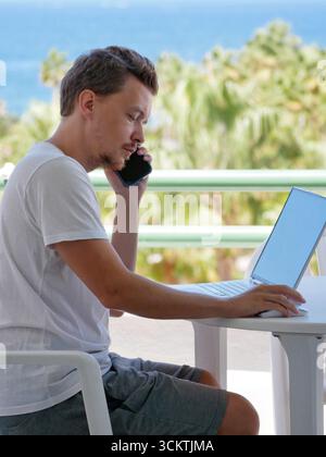 Man working remotely by the sea, speaking on smartphone while using a laptop outdoors. Concepts like remote work, workation, and location independence Stock Photo