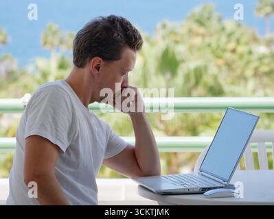 A focused and thoughtful young man working on a laptop outdoors with a beautiful ocean and palm tree background. Themes of freedom, flexibility, and m Stock Photo