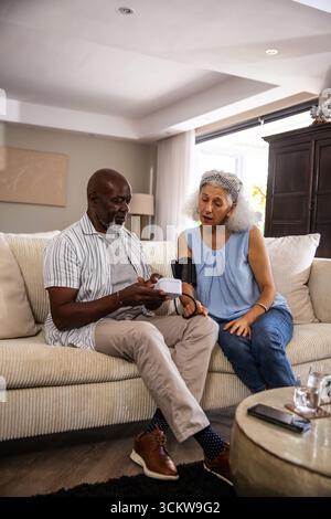 Diverse senior couple sitting in living room monitoring blood pressure using digital cuff Stock Photo