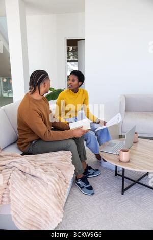 Diverse couple reviewing documents while sitting on sofa at home with laptop and coffee mugs Stock Photo