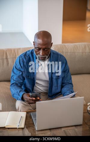 African american senior man using laptop and smartphone, reviewing papers and notebook at home Stock Photo