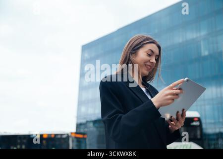 Professional woman engaged with a tablet in an urban environment, surrounded by modern architecture, highlighting technology and connectivity Stock Photo