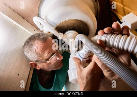 Kitchen sink trap with dishwasher connection under basin, plumber installs new trap and secures waste outlet by tightening plastic nut. Stock Photo