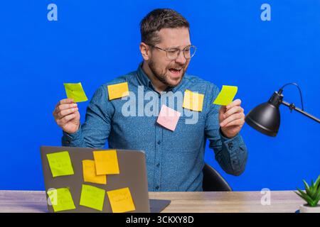 Exhausted man sitting at table with pasted sticker notes use laptop computer having concentration problem creative crisis. Nervous adult guy confused by big routine work loading on blue background Stock Photo