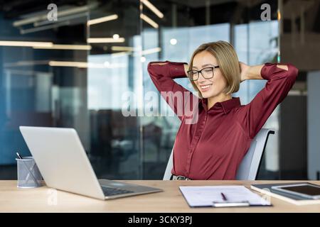 Happy businesswoman is relaxing at her desk with hands behind her head, smiling and looking at her laptop after finishing work in a modern office Stock Photo