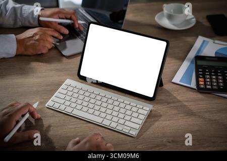 Digital tablet mockup concept: iPad with blank white screen placed on wooden office desk, alongside keyboard, stylus, laptop, calculator, and coffee c Stock Photo
