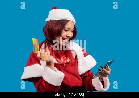 Happy young woman wearing santa claus costume using smartphone and credit card, shopping online for christmas gifts against a blue background Stock Photo