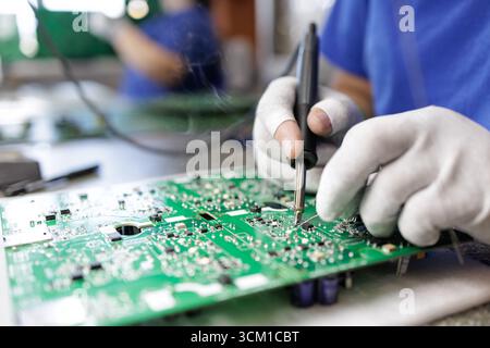 Closeup of ESD gloved worker soldering parts on a green PCB at an electronics assembly workstation in a modern factory Stock Photo