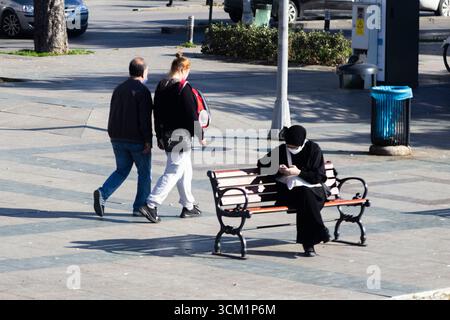 People in face masks walk past a woman sitting on a park bench, public space, daily life, pandemic Stock Photo