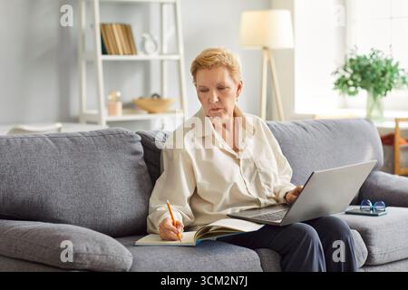 Mature woman is focused on work, taking notes in notebook while using her laptop. Stock Photo