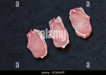 Top view of three fresh raw pork chops placed on a dark textured surface with scattered flour, peppercorns, garlic cloves, bay leaves, onion, and herb Stock Photo