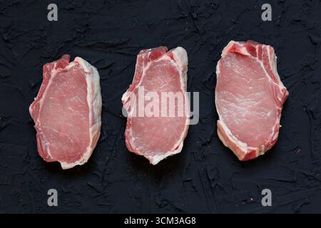Top view of three fresh raw pork chops placed on a dark textured surface with scattered flour, peppercorns, garlic cloves, bay leaves, onion, and herb Stock Photo
