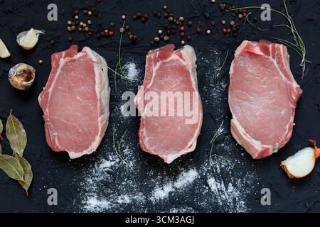 Top view of three fresh raw pork chops placed on a dark textured surface with scattered flour, peppercorns, garlic cloves, bay leaves, onion, and herb Stock Photo